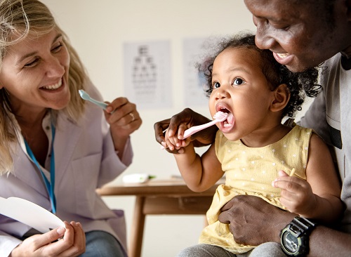 A little girl is in the doctors office with a doctor and caregiver, demonstrating teeth brushing
