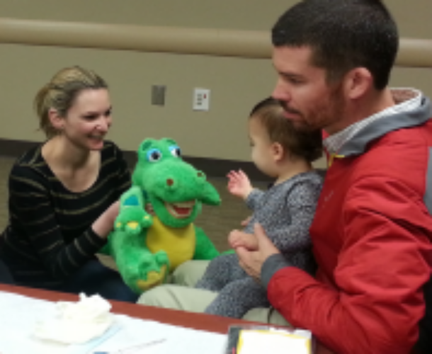 A father with his infant sitting on his lap looking at a woman who is holding a dinosaur puppet.